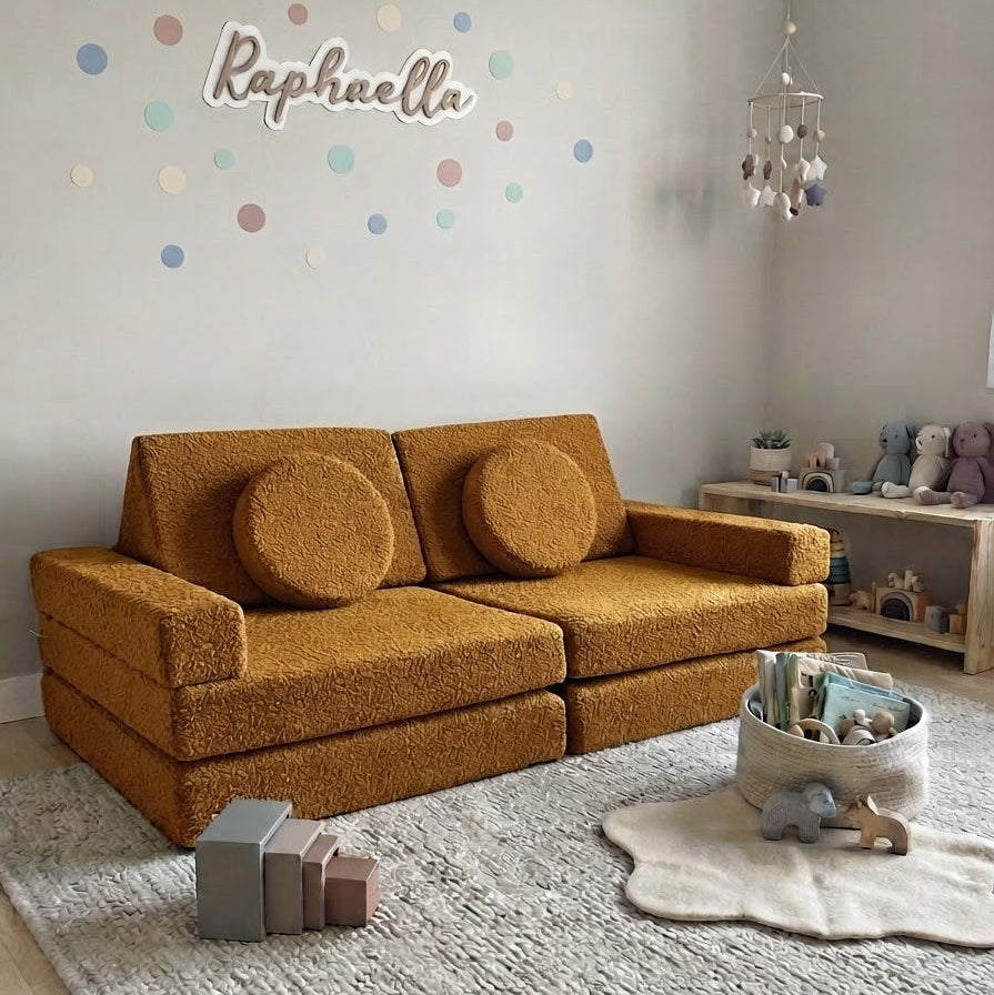 A mustard yellow textured boucle sofa in a styled children's room, featuring a matching round throw pillow, a light gray rug, and a wall with colorful polka dots and the name 'Raphaella'.