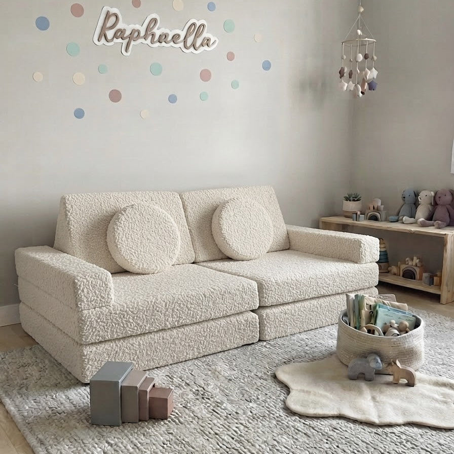 A cream bouclé two-seater sofa with two matching round throw pillows in a styled nursery or children's room. The room features a textured light gray rug, a wooden toy shelf, and a wall decorated with pastel polka dots and the name 'Raphaella'.