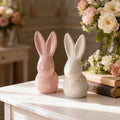 Two ceramic rabbit figurines on a white table with floral arrangements in the background.