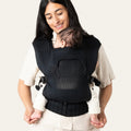 Woman carrying a baby in a black ergonomic carrier against a plain background
