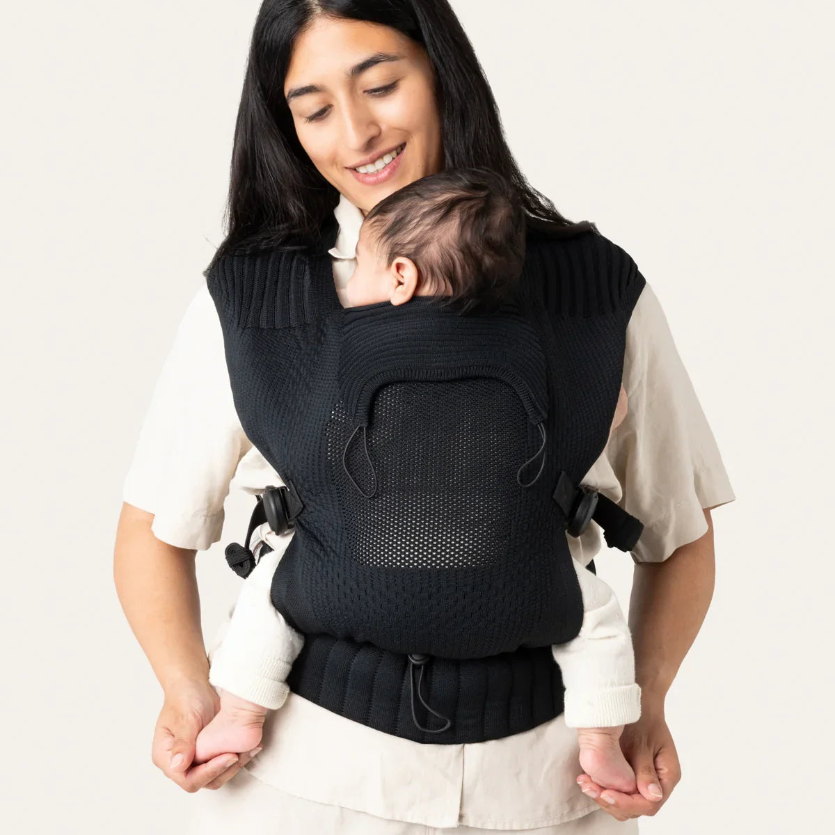 Woman carrying a baby in a black ergonomic carrier against a plain background
