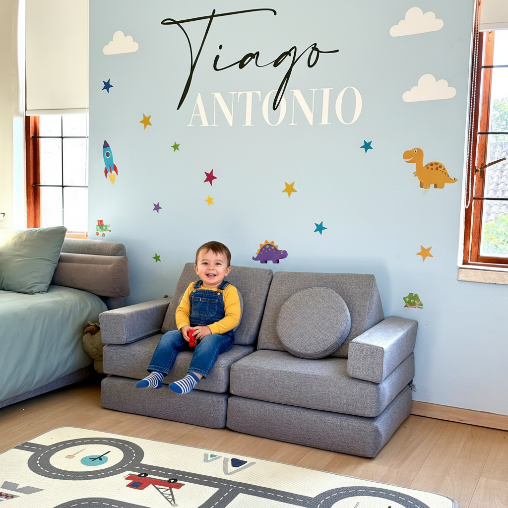 A child sits on a gray modular sofa in a bright room with a light blue wall decorated with the name 'Tiago ANTONIO' in script and block letters, along with space-themed decals including stars, clouds, a rocket, and a dinosaur.