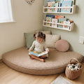 A young child sits cross-legged on a round, low-profile beige floor cushion in a cozy room corner, reading a book. The cushion is styled with neutral-toned pillows, next to a woven basket of stuffed toys, with light wood bookshelves holding children's books on the wall above.