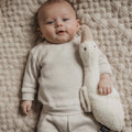 Baby in a white outfit holding a plush toy on a textured beige blanket
