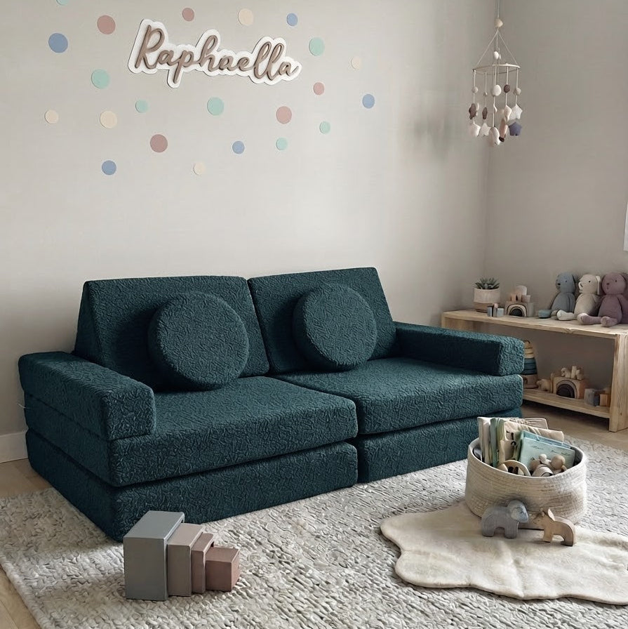 A dark teal textured fabric sofa in a styled children's room, featuring a light gray rug, a wooden toy shelf, and a personalized wall decal that reads 'Raphaella' with pastel polka dots.