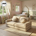 A beige upholstered two-seater sofa with two round matching throw pillows, shown in a styled children's bedroom. The room features soft pink walls, a house-shaped bed, a small bookshelf, and warm string lights.