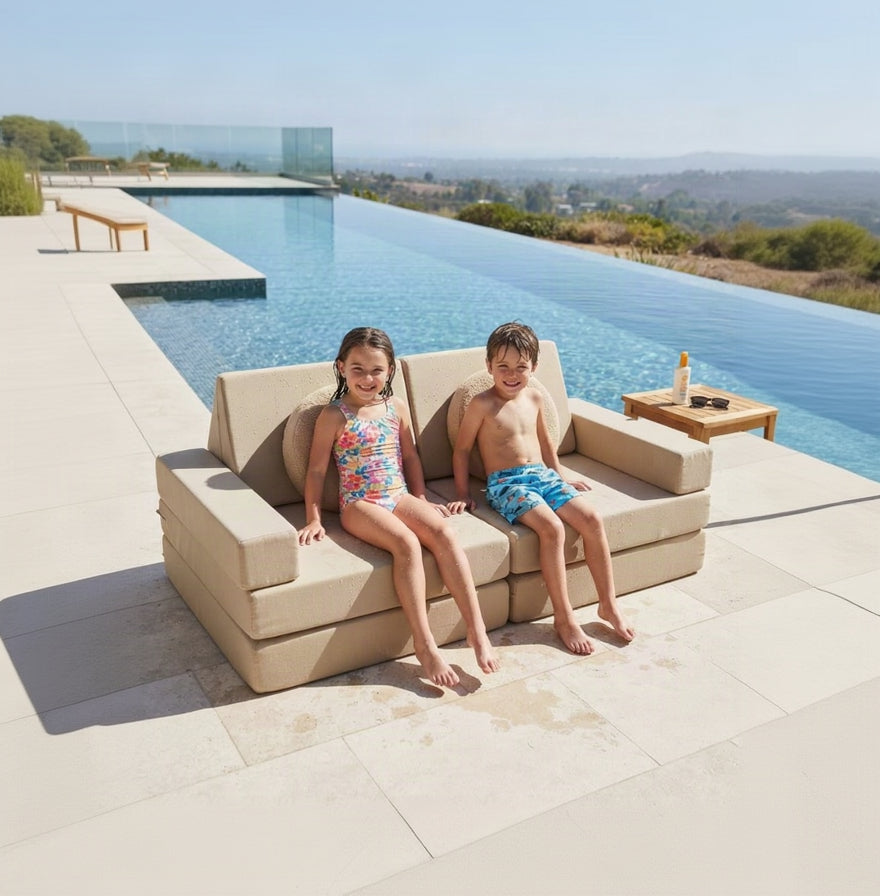 Two children sitting on a beige outdoor sectional sofa next to an infinity pool, with a scenic landscape in the background.