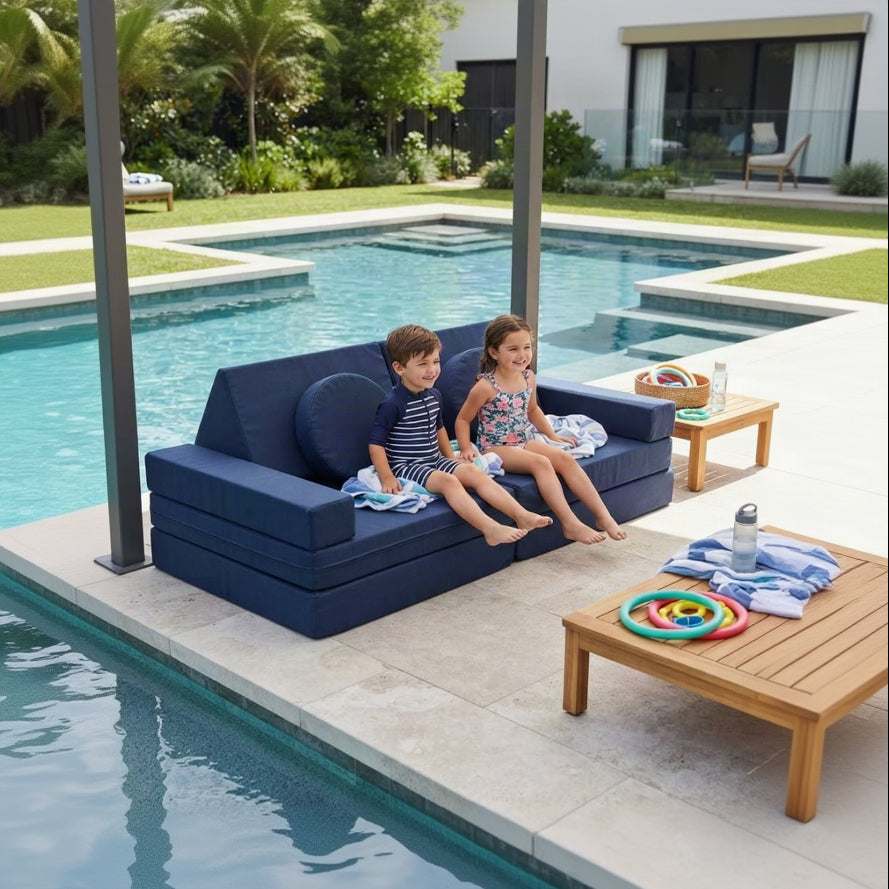 Two children sitting on a navy blue outdoor poolside sofa next to a swimming pool, with a wooden lounge table and poolside setting in the background.