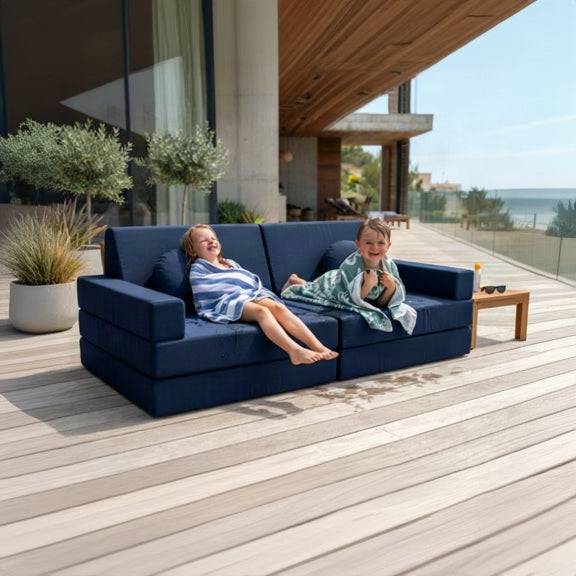 A navy blue outdoor sectional sofa on a wooden deck, with two children reclining on it and a beach and ocean visible in the background.