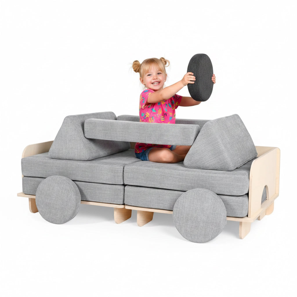 A child sits inside a car-shaped modular play sofa made of light wood and gray fabric, holding a round gray cushion against a clean white background.