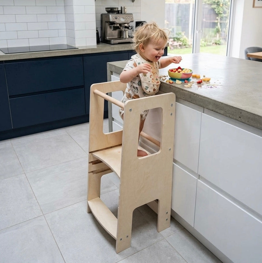 Child in a wooden learning tower at a kitchen counter with a modern kitchen setting.