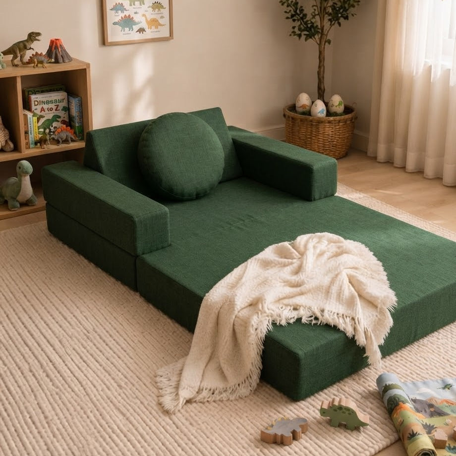 A dark green modular foam daybed with a matching round cushion and a cream throw blanket, styled in a child's bedroom with dinosaur-themed decor, a bookshelf, and a potted tree.