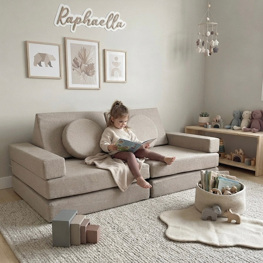 A child sits on a light beige upholstered sofa in a softly styled nursery or playroom, reading a book. The room has pale walls, framed wall art, a textured rug, toys, and a hanging mobile.