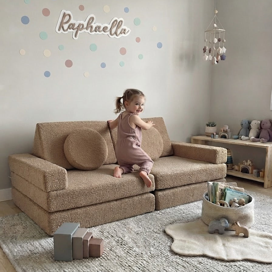 A toddler girl in a pink outfit sits on a beige boucle sofa in a styled nursery room. The room features a textured light grey rug, a wooden toy shelf, and a personalised wall decal.