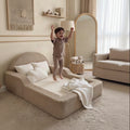 A child wearing a matching beige pajama set jumps on a low upholstered bed in a softly styled bedroom. The room has warm cream walls, a large floor mirror, a rug, and neutral-toned furniture.