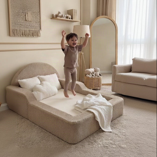 A child wearing a matching beige pajama set jumps on a low upholstered bed in a softly styled bedroom. The room has warm cream walls, a large floor mirror, a rug, and neutral-toned furniture.