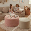 A cozy children's living room scene with two young girls sitting on a beige sofa, surrounded by soft home decor including a pink corduroy round ottoman, a cream cylindrical ottoman, a beige shag rug, and a basket of teddy bears.