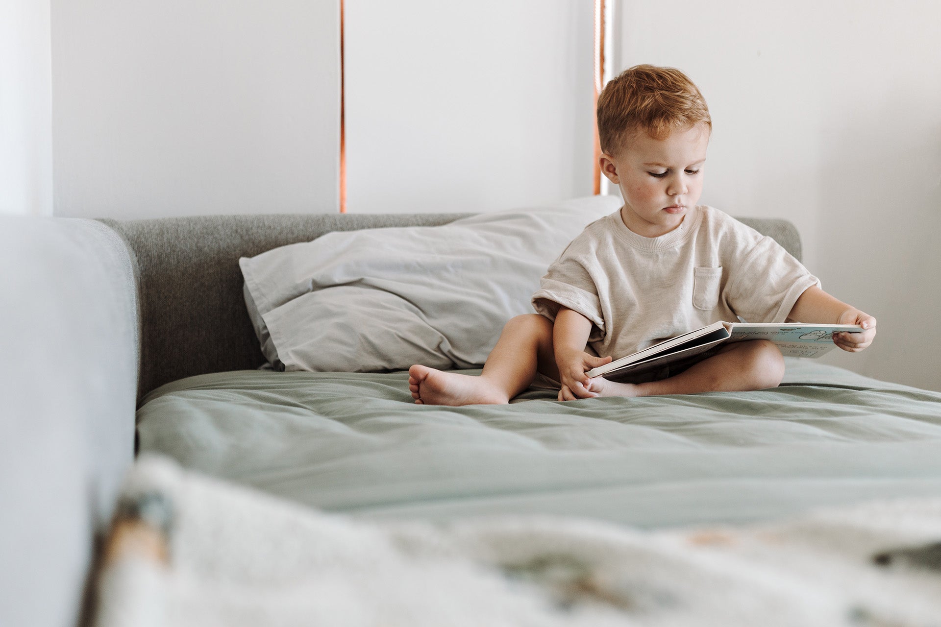 Child sitting on a bed reading a book in a cozy bedroom.